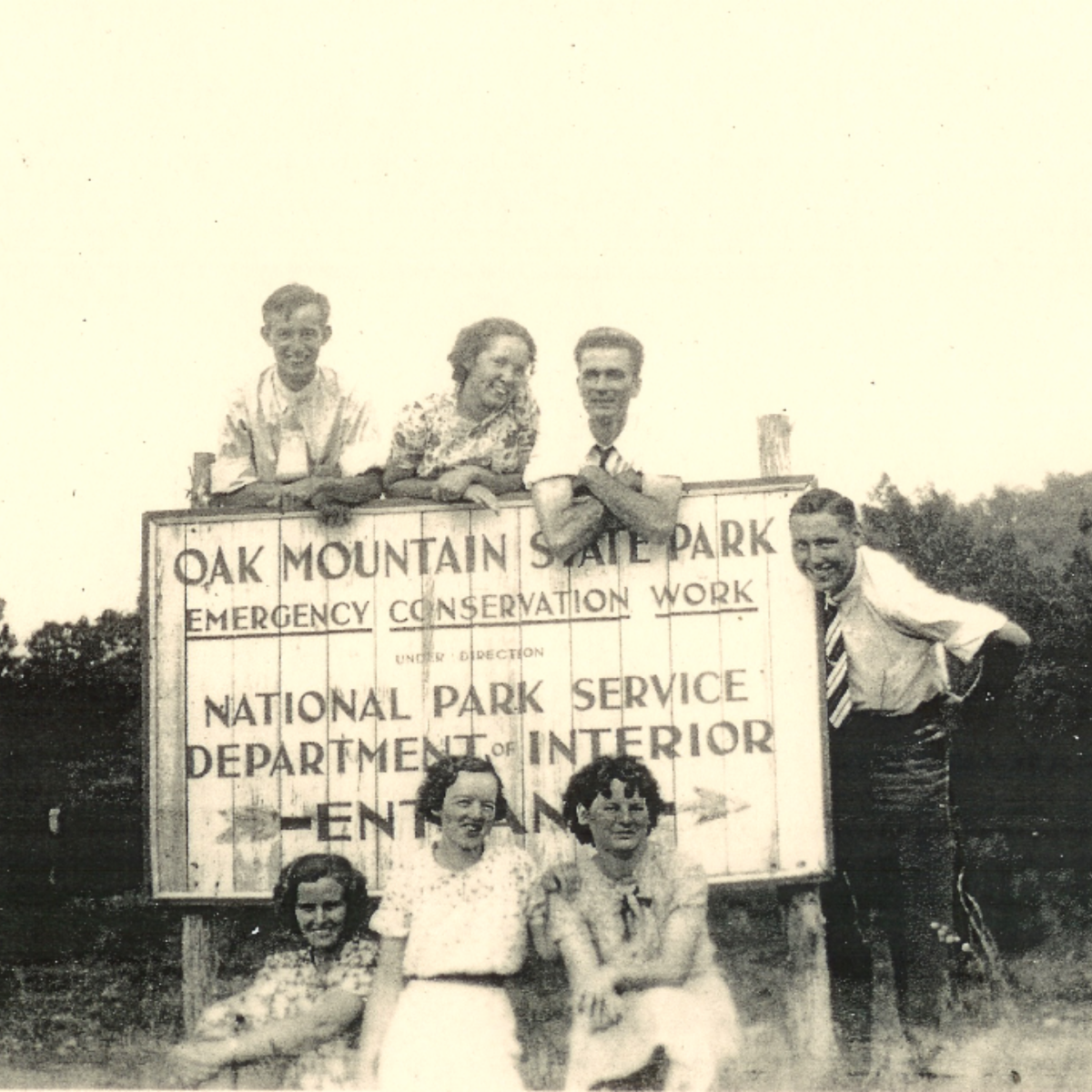 Yellowed black and white photo of 4 young women and 3 young men around a sign for Oak Mountain State Park 